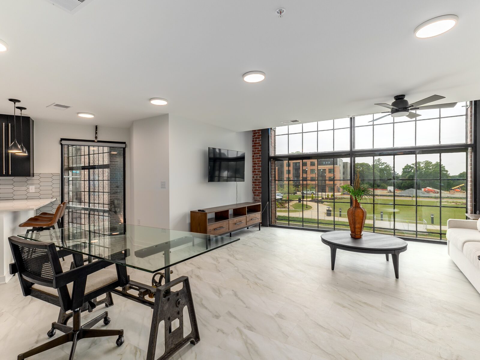 Open-plan living room with exposed brick walls, floor-to-ceiling factory windows, and hardwood floors inside a Power House loft apartment in Rock Hill, SC